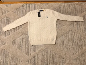 Vit  tröja Polo Ralph Lauren - Snygg vit kabelstickad tröja från Polo Ralph Lauren med klassisk rund hals och svart broderad logga på bröstet. Tröjan har långärmad passform och ribbade muddar vid ärmslut och nederkant. Perfekt för dig som gillar stilren och tidlös design.