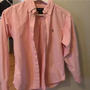 Rosa skjorta från Ralph Lauren - Klassisk rosa skjorta från Ralph Lauren med button down-krage och broderad logga på bröstet. Skjortan har långa ärmar och knäppning framtill. Passar dig som är runt 165. Pris kan diskuteras