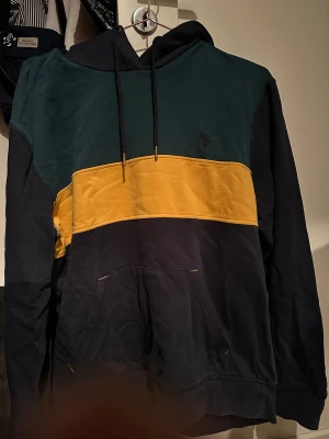 hoodie från U.S. Polo Assn. - Snygg hoodie från U.S. Polo Assn. i storlek M med färgblock i mörkgrön, gul och svart. Tröjan har huva med snörning och en stor magficka. Perfekt för chill dagar och streetstyle. Materialet är mjuk bomull som känns skönt mot huden.