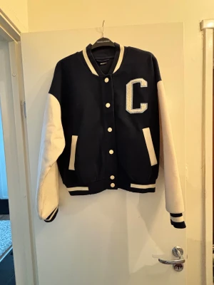 Blå och vit bomberjacka California - Snygg bomberjacka i mörkblått med vita ärmar och detaljer. Stor broderad bokstav C på bröstet och texten CALIFORNIA på ryggen. Jackan har ribbade muddar, knappar framtill och två fickor.