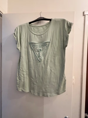Ljusgrön t-shirt från Guess - Snygg ljusgrön t-shirt från Guess med det klassiska logotrycket i form av en triangel framtill. T-shirten har korta ärmar och rund hals. Perfekt för dig som gillar stilrena och trendiga plagg med tydlig branding.