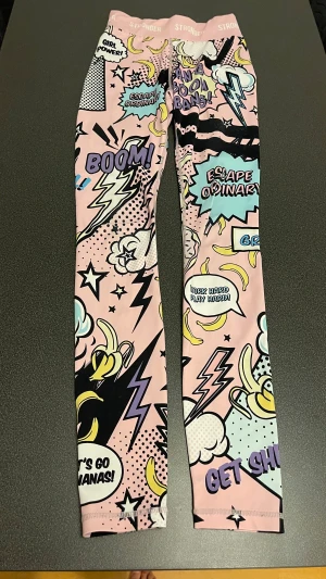 Stronger leggings med pop art-mönster - Säljer ett par färgglada leggings från Stronger med pop art-inspirerat tryck i rosa, blått, svart och gult. De har hög midja och coola motiv som blixtar, bananer och peppande texter. Perfekta för träning eller när du vill sticka ut.