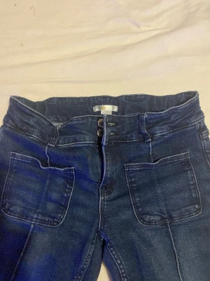 Mörkblå jeans från Gina Tricot - Snygga mörkblå jeans från Gina Tricot med raka ben och hög midja. Jeansen har två stora framfickor och dubbla knappar i midjan för extra cool detalj. Materialet är klassisk jeans med lite stretch för skön passform.