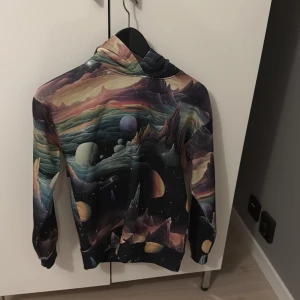 Galaxy hoodie med färgstarkt tryck - Unik hoodie med ett färgstarkt galaxmotiv som täcker hela tröjan. Mönstret har planeter, berg och rymdlandskap i nyanser av svart, lila, blått, beige och orange. Tröjan har huva och långa ärmar, perfekt för dig som gillar coola prints och vill sticka ut.