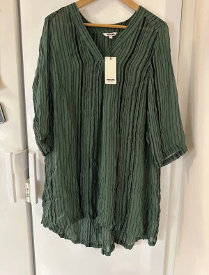 Ny tunika från Indiska st. S - Säljer en ny  luftig grön tunika med svarta ränder från Indiska med svarta ränder och v-ringad hals. Tunikan har trekvartsärmar och är tillverkad i ett lätt, transparent materia. Den har en avslappnad passform.  Svårt med exakt färg på bilderna. Skulle säga att färgen är något ljusare än bilderna visar.  100% viskos   Modell: Grishma tunic 221