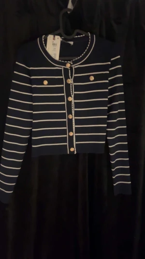 Randig kofta från Holly & Whyte XS - Supersnygg marinblå och vit randig kofta från Holly & Whyte. Croppad modell med guldfärgade knappar framtill och dekorativa fickor. Stickad material med långärmad passform och fina detaljer runt halsen. Perfekt för dig som gillar klassisk stil med en twist.