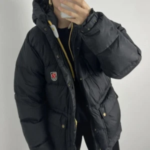 Svart dunjacka från Fjällräven - Säljer en svart dunjacka från Fjällräven med huva och dragkedja. Klassisk Fjällräven-logga på bröstet. Perfekt för kalla dagar och riktigt snygg streetstil. Har några få defekter som är ihop sydda och saknar en knapp men annars helt i funktion.