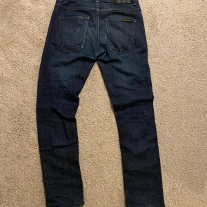 Mörkblå jeans från Nudie Jeans - Snygga mörkblå jeans från Nudie Jeans med klassisk femficksdesign och subtila kontrastsömmar på bakfickorna. Jeansen har rak passform och är tillverkade i slitstarkt bomullsdenim. Perfekta för dig som gillar stilrena och tidlösa jeans.