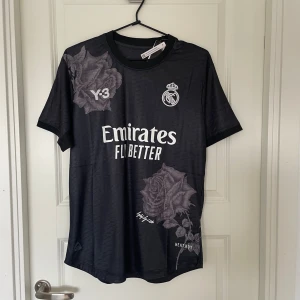 Svart Real Madrid Y-3 x Adidas fotbollströja - Snygg svart Real Madrid fotbollströja från Y-3 x Adidas med vita tryck och stora gråa rosor som detaljer. Kortärmad modell i slim fit med rund hals och Heat.RDY-material som andas. Perfekt för dig som vill sticka ut på planen eller i stan.