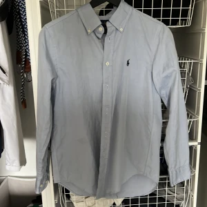 Ljusblå skjorta från Ralph Lauren - Snygg ljusblå skjorta från Ralph Lauren med klassisk button down-krage och broderad logga på bröstet. Skjortan har lång ärm, knappar framtill och är i bomull. Perfekt för en clean och stilren look.