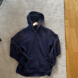 Marinblå hoodie från Polo Ralph Lauren - Säljer en marinblå hoodie från Polo Ralph Lauren med dragkedja och huva. Klassisk röd broderad logga på bröstet och kängurufickor framtill. Perfekt för chill dagar och snygg till jeans eller joggers.