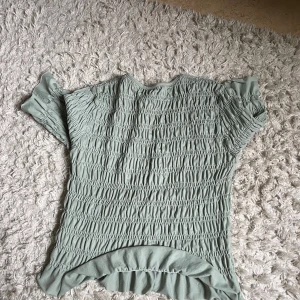 Ljusgrön blus med smock och volang - Supersöt ljusgrön blus med smock över hela plagget och volangdetalj längst ner. Blusen har rund halsringning och trekvartsärm med lätt puff. Perfekt för dig som gillar en feminin och trendig look.