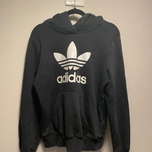 Svart Adidas hoodie med stor logga - Svart hoodie från Adidas med stor vit Trefoil-logga och text på bröstet. Klassisk känguruficka framtill, huva och ribbade muddar. Mjuk insida och avslappnad passform, perfekt för chill eller streetwear.
