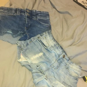 Två par jeansshorts i blå toner - Säljer två par snygga jeansshorts i olika blå nyanser. Ena paret är mörkblå med råa kanter och dubbla knappar fram, medan det andra är ljusblått med slitningar och fransiga detaljer. Båda har klassiska fickor och bälteshällor, perfekta för sommarens chill.