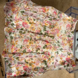 Blommig kortkjol från Primark - Supersöt kortkjol från Primark med blommigt mönster i rosa, gult och grönt på vit botten. Kjolen har volangdetaljer och är tillverkad i ett lätt, luftigt material med innerfoder och dragkedja bak. Perfekt för vår och sommar!