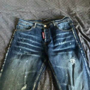 Dsquared2 blå slitna jeans - Riktigt snygga blå jeans från Dsquared2 med slitningar och trasiga detaljer på benen. Klassisk femficksmodell med tydliga kontrastsömmar och röd Dsquared2-tag vid fickan. Jeansen har en cool, streetig vibe och är tillverkade i jeansmaterial. har haft på mig dom ett få gånger så dom är i nyskick
