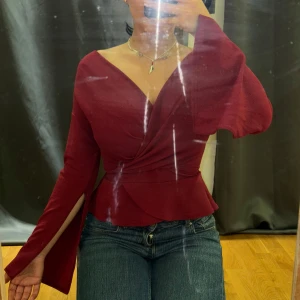 Vinröd omlottopp från Boohoo - Säljer en vinröd omlottopp från Boohoo med v-ringning och långa trumpetärmar. Toppen har knyt i midjan och en figurnära passform som framhäver midjan. Materialet är mjukt och lätt, perfekt för att skapa en snygg siluett.