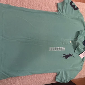 Grön Polo Ralph Lauren pikétröja - Snygg grön pikétröja från Polo Ralph Lauren i slim fit-modell. Klassisk krage, två knappar och stor marinblå logga broderad på bröstet. Tillverkad i bomull och har korta ärmar med siffra på ena ärmen. Perfekt för en clean och sportig look. Den passar för ålder 14-16 L och är också ny som ni ser priset är kvar o allt helt oanvänd