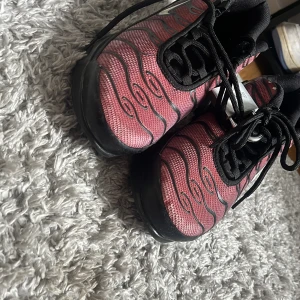 Nike Air Max Plus Tn rosa och svarta - Nike Air Max Plus Tn sneakers i rosa och svart med vågigt mönster och silvriga detaljer. Skorna har svart sula, snörning och mesh-överdel för en sportig vibe. Ikonisk Tn Air-logga på hälen och klassisk Swoosh på sidan. Perfekta för dig som vill sticka ut.