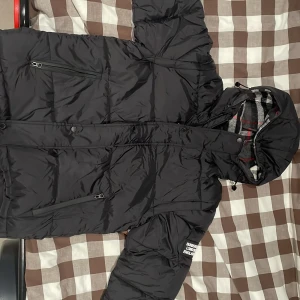 Svart pufferjacka från Burberry - Svart pufferjacka från Burberry med avtagbara ärmar och huva fodrad i klassiskt rutigt mönster. Jackan har dragkedja, knappar och flera fickor med dragkedja. Perfekt för kalla dagar och riktigt snygg streetstil.  Kan diskutera priset 