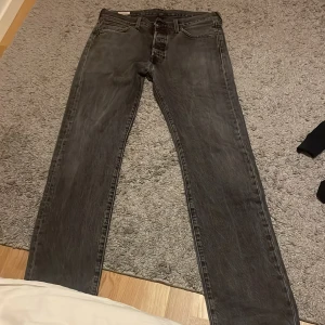Levi's 501 svarta jeans 32/34 - Klassiska svarta Levi's 501 jeans i rak modell. Jeansen har fem fickor, knappgylf och den ikoniska läderpatchen bak i midjan. Perfekt för dig som gillar en tidlös och avslappnad stil. Färgtonen är mörkgråsvart och materialet är robust denim.