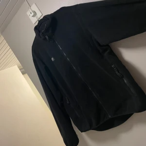 Svart fleecejacka från Helly Hansen - Svart fleecejacka från Helly Hansen med hög krage och hel dragkedja framtill. Jackan har två sidofickor med dragkedja och en liten HH-logga på bröstet. Perfekt för kyliga dagar och enkel att matcha med allt.