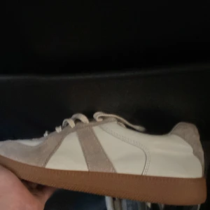 Maison margiela  - Snygga sneakers i vitt och beige med bruna gummisulor. Skorna har ovandel i skinn och mocka, med klassisk snörning och låg profil. Perfekta för dig som gillar stilrena och tidlösa sneakers med en touch av retro.