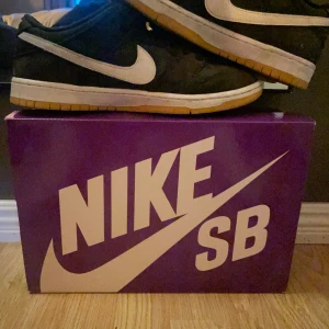 Svarta Nike SB Dunk sneakers - Säljer ett par svarta Nike SB Dunk sneakers med vit swoosh. Skorna har klassisk låg profil och är perfekta för dig som gillar streetwear eller skate. Kommer med originalkartong i lila med stor Nike SB-logga.