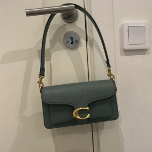 Coach väska - Helt oanvänd grön (sage) axelväska från Coach i äkta skinn med justerbar rem och ikoniskt C-spänne i guld. Väskan har ett stilrent, rektangulärt format och flera fack inuti, inklusive ett med dragkedja. Perfekt för dig som vill ha en trendig och praktisk väska.