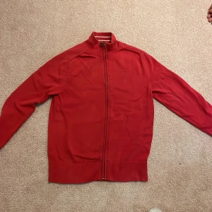 Röd kofta från Tommy Hilfiger - Röd kofta från Tommy Hilfiger med hel dragkedja och ribbade muddar. Klassisk design med liten broderad logga på bröstet och ståkrage med vita ränder på insidan. Perfekt för dig som gillar stilrena och snygga plagg.