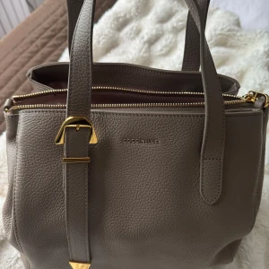 Beige handväska från Coccinelle i skinn - Snygg beige handväska från Coccinelle i mjukt, kornigt skinn med guldfärgade metalldetaljer och dubbla handtag. Väskan har dragkedja upptill och stilren design med diskret logga framtill. Perfekt storlek för vardagens essentials.
