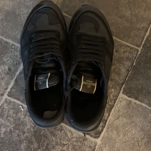 Svarta Valentino Rockrunner sneakers - Säljer ett par svarta Valentino Rockrunner sneakers med kamouflagemönster och detaljer i mocka och skinn. Skorna har snörning och en platt sula med nitar på hälen. Perfekta för dig som gillar exklusiva och stilrena sneakers.
