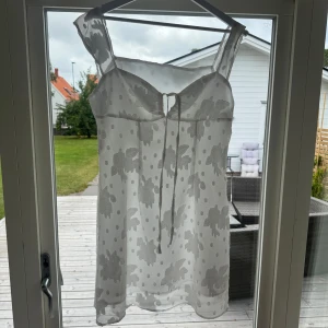 Vit blommig kortklänning med snörning - Supersöt vit kortklänning med blommigt och prickigt mönster. Klänningen har tunna axelband, lätt transparent tyg och snörning framtill vid bysten. Perfekt för varma dagar och har en flowy passform som ger en chill vibe.