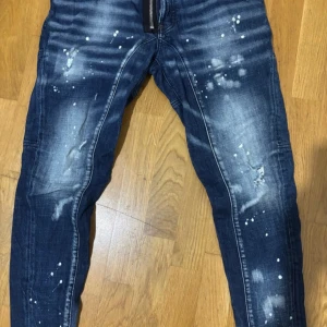 Mörkblå DSQUARED2 jeans med splatter - Snygga mörkblå jeans från DSQUARED2 med coolt splattermönster och slitningar. Modellen har raka ben med förstärkta sömmar vid knäna, klassiska fem fickor och dragkedja med DSQUARED2-logga. Perfekt för dig som gillar streetstyle och detaljer. Fräscha i stilen, passar med allt möjligt. Bekväma i långa dagar samt korta, inte alls för lösa eller thigta riktigt perfekt.