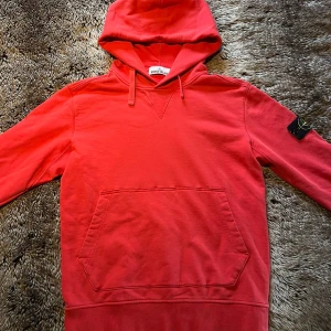 Röd hoodie från Stone Island - Säljer en röd hoodie från Stone Island med klassisk patch på vänster ärm. är tillverkad i mjuk bomull. Perfekt för dig som gillar lite färg.