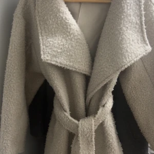 Beige teddyjacka med bälte - Mysig beige teddyjacka med bred krage och knytskärp i midjan. Jackan har en oversized look och är perfekt för kyliga dagar. Materialet är fluffigt och ger en trendig vibe.