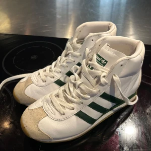 Adidas vita och gröna high-top sneakers - Säljer ett par klassiska Adidas high-top sneakers i vitt skinn med gröna ränder och detaljer. Skorna har snörning, beige mockadetaljer på tån och hälen samt gummisula. Perfekta för dig som gillar retrostil och vill sticka ut med en ikonisk look.