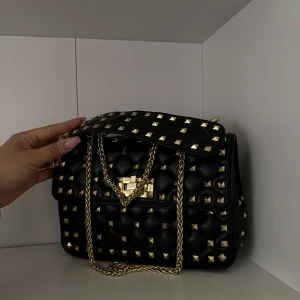 Valentino liknande - Säljer en svart axelväska med quiltad design och guldiga nitar över hela väskan. Väskan har en snygg guldfärgad kedja som axelrem och ett matchande lås framtill. 