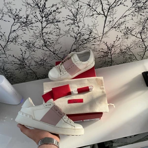 Valentino Open sneakers vit/rosa - Säljer ett par Valentino Open sneakers i vitt skinn med bred rosa rem över mitten och ikoniska nitar på hälen. Klassisk rund tå, platt sula och vita snören. Kommer med originalkartong, extra skosnören och dustbag. Perfekta för dig som gillar lyxiga detaljer. Medföljer: Box, extraskosnören, kort från valentino, dustbag