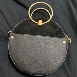Rund svart handväska med guldringar - Snygg rund handväska i svart skinnimitation och mockadetalj upptill. Väskan har två stora guldfärgade ringar som handtag och stängs med dragkedja. Perfekt för dig som vill sticka ut med en unik accessoar. Enkel men ändå trendig design.