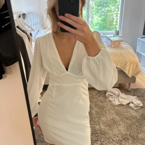 Vit kort omlottklänning med v-ringning - Snygg vit kort klänning med djup v-ringning och långa puffärmar. Klänningen har en omlottliknande överdel och markerad midja som ger en smickrande siluett. Perfekt för dig som gillar stilrena och trendiga plagg.