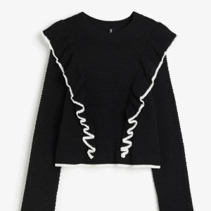 Svart stickad tröja med volangdetaljer - Svart stickad tröja med långa ärmar och vita volangdetaljer längs axlar och framsida. Storlek M