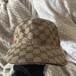 Gucci keps med GG-mönster, beige - Säljer en beige keps från Gucci med klassiskt GG-monogram över hela kepsen. Den har justerbar rem bak i mörkbrunt skinn och detaljer i grönt och rött på sidorna. Materialet är bomull och kepsen är tillverkad i Italien. Perfekt för dig som vill ha en lyxig streetstyle.