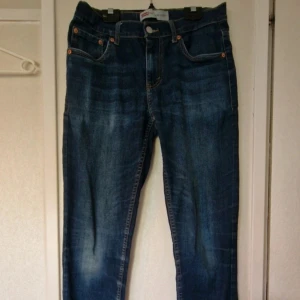 Levi's 512 Slim Taper jeans blå - Snygga Levi's 512 Slim Taper jeans i klassisk blå denim med orange sömmar och femficksdesign. Jeansen har en smal passform som smalnar av nedtill och är tillverkade i slitstarkt bomullsjeans. Perfekta för dig som gillar en modern och stilren look. Det är ett hål i höger ficka och märket bak har börjat gå upp. Stl 16A