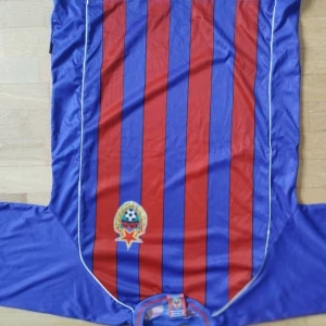 CSKA Moskva kortärmad fotbollströja XL  - Retro läkortärmad fotbollströja från CSKA Moskva med vertikala blå och röda ränder, klubbmärke på bröstet och nummer 8 med rysk text på ryggen. Tillverkad i glansigt syntetmaterial, klassisk passform och vita detaljer längs sidorna.