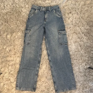Blå cargojeans med raka ben - Säljer ett par ljusblå cargojeans med raka ben och stora fickor på sidorna. Jeansen har klassisk dragkedja och knapp framtill samt bälteshällor. Perfekta för dig som gillar streetstyle och vill ha plats för allt i fickorna. Aldrig använd 