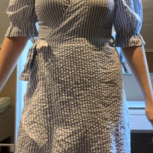 Randig omlottklänning med puffärm - Superfin kort omlottklänning med blåvita ränder och puffärmar. Klänningen har knyt i midjan och v-ringad hals. Tillverkad i ett lätt bomullstyg som känns perfekt för varmare dagar. Snygg passform och trendig look!