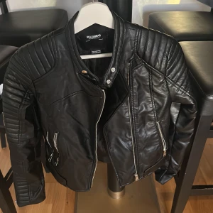 Svart skinnjacka från Pull&Bear XS - Snygg svart skinnjacka från Pull&Bear i storlek XS. Jackan har coola quiltade detaljer på axlar och ärmar, flera dragkedjor framtill och en klassisk bikerstil. Perfekt för dig som vill ha en edgy look med stilrena detaljer.