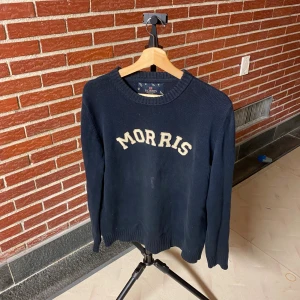 Mörkblå stickad tröja från Morris - Snygg mörkblå stickad tröja från Morris med krämvit broderad logga framtill. Klassisk rund halsringning och ribbade muddar vid ärmslut och nederkant. Perfekt för dig som gillar stilren och tidlös design.