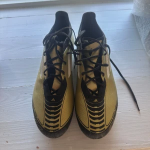 Adidas Messi fotbollsskor guld/svart - Säljer ett par Adidas Messi fotbollsskor i guld och svart med vita detaljer. Skorna har snörning, platt sula med dobbar och en snygg grafisk design på ovansidan. Perfekt för dig som vill sticka ut på planen med stilrena och sportiga skor.